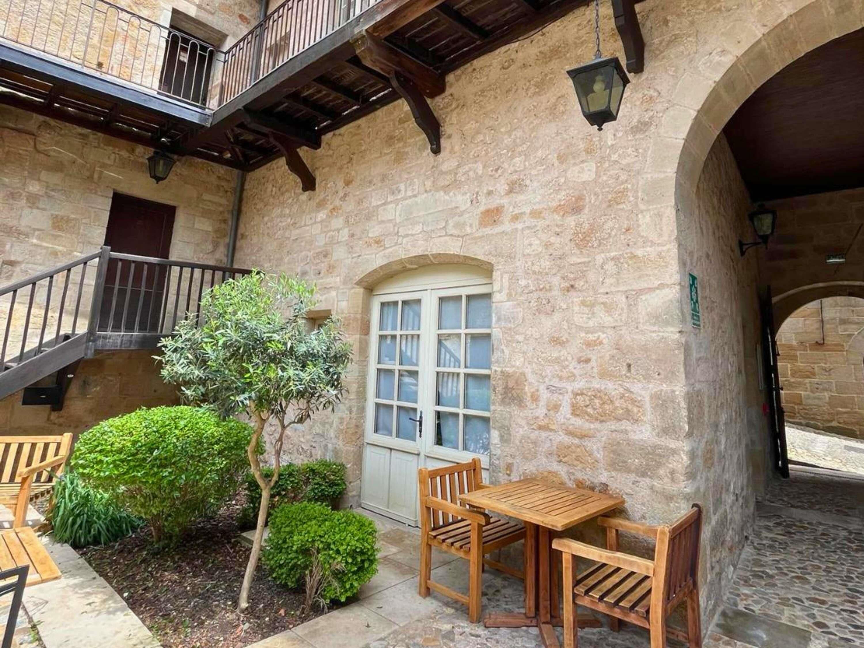 Un edificio de piedra con un balcón de madera, una puerta y un pequeño jardín con bancos y una mesa.