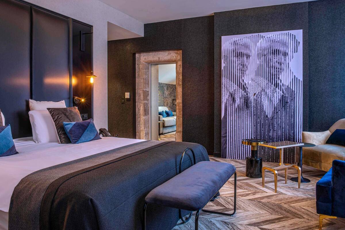 Ein modernes Hotelzimmer mit einem großen Bett, einer Sitzecke inklusive Tisch und Stühlen sowie einem großen abstrakten Kunstwerk an der Wand.