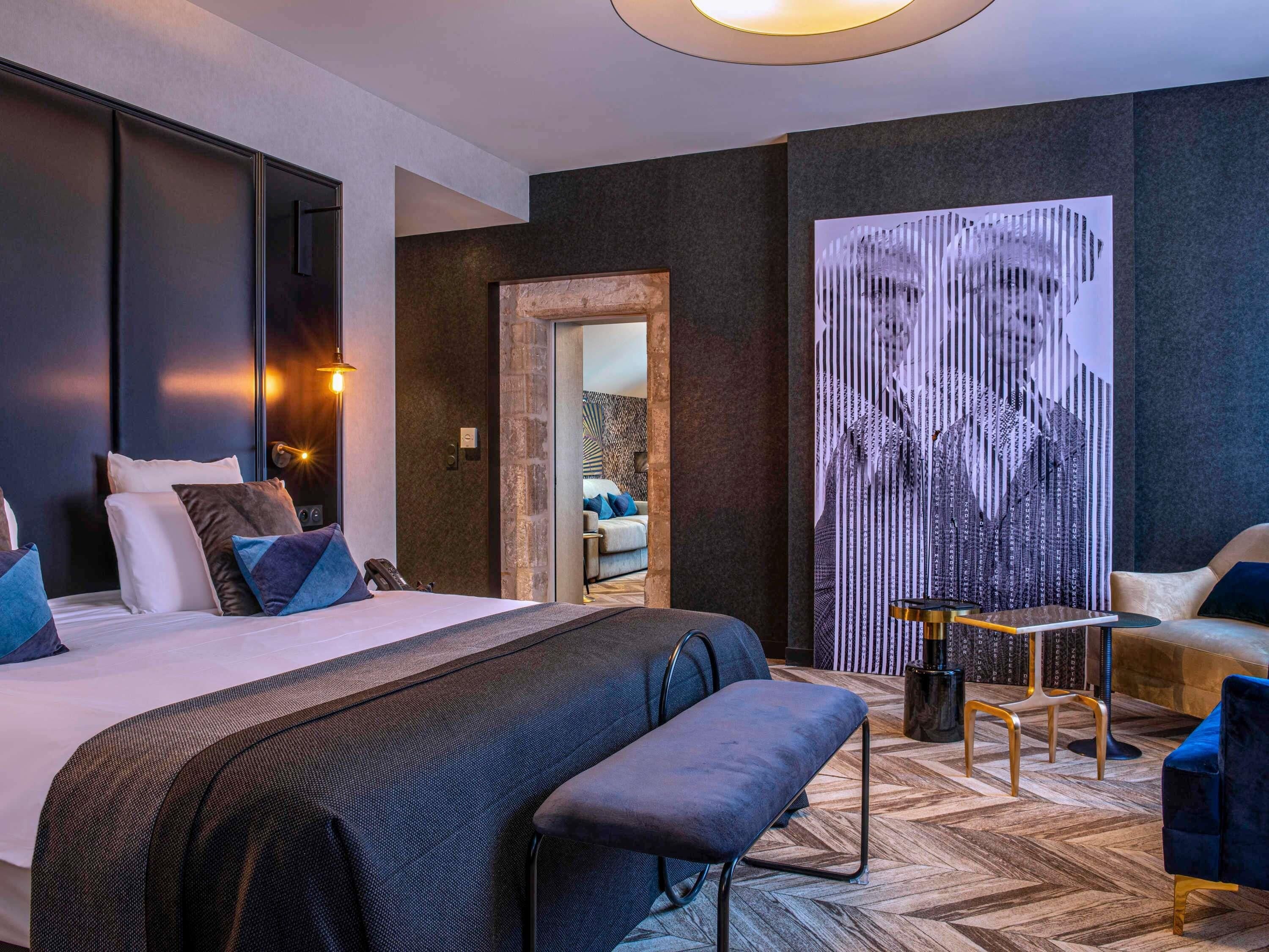 Ett modernt hotellrum med en stor säng, en sittgrupp med bord och stolar samt ett stort, abstrakt konstverk på väggen.
