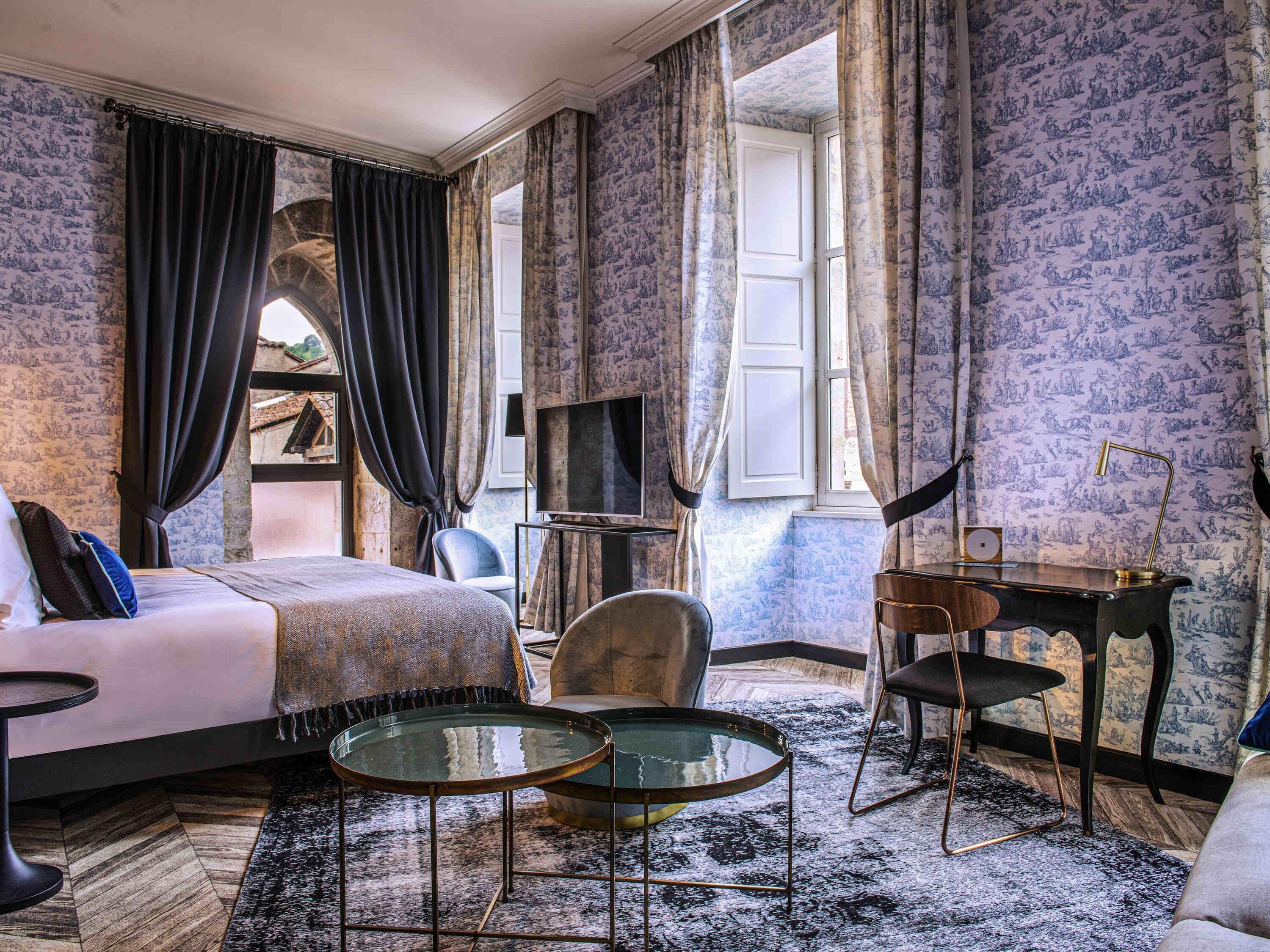 Une chambre d’hôtel avec un lit, deux chaises, une petite table, un bureau, une télévision et un papier peint à motifs.