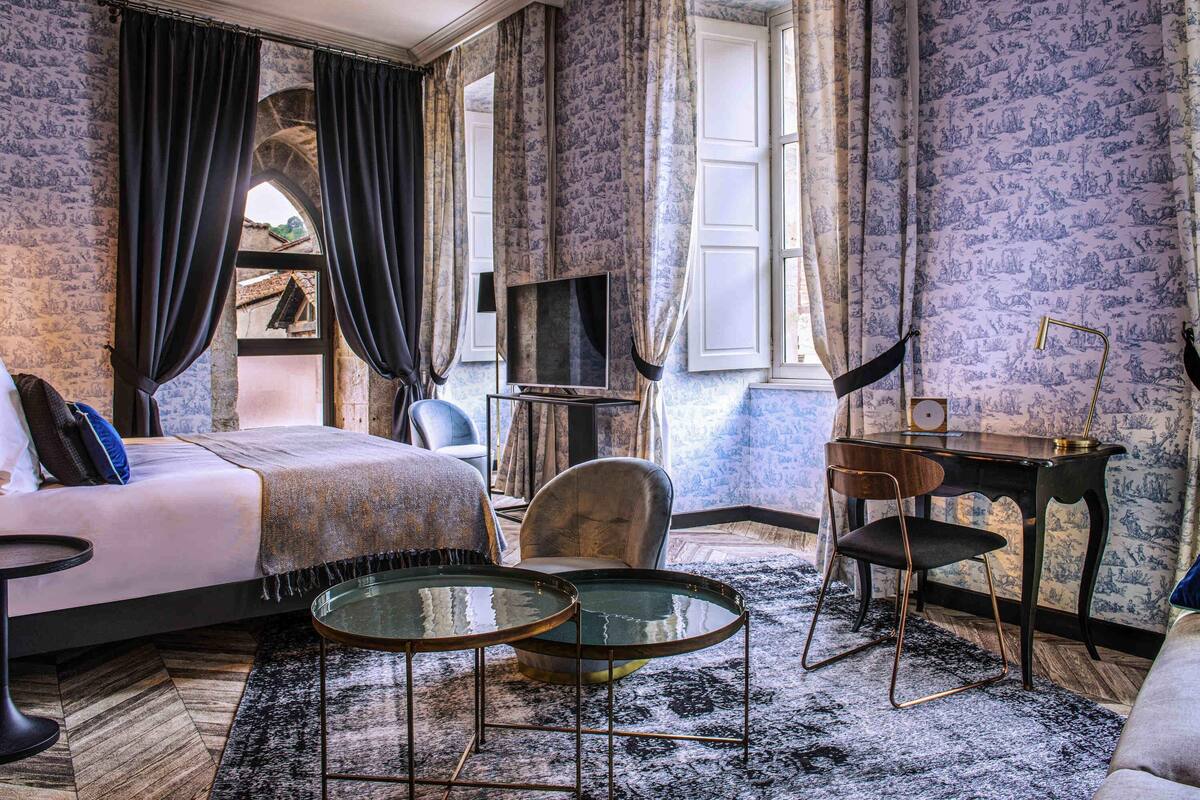 Ein Hotelzimmer mit einem Bett, zwei Stühlen, einem kleinen Tisch, einem Schreibtisch, einem Fernseher und Wandtapete mit Muster.