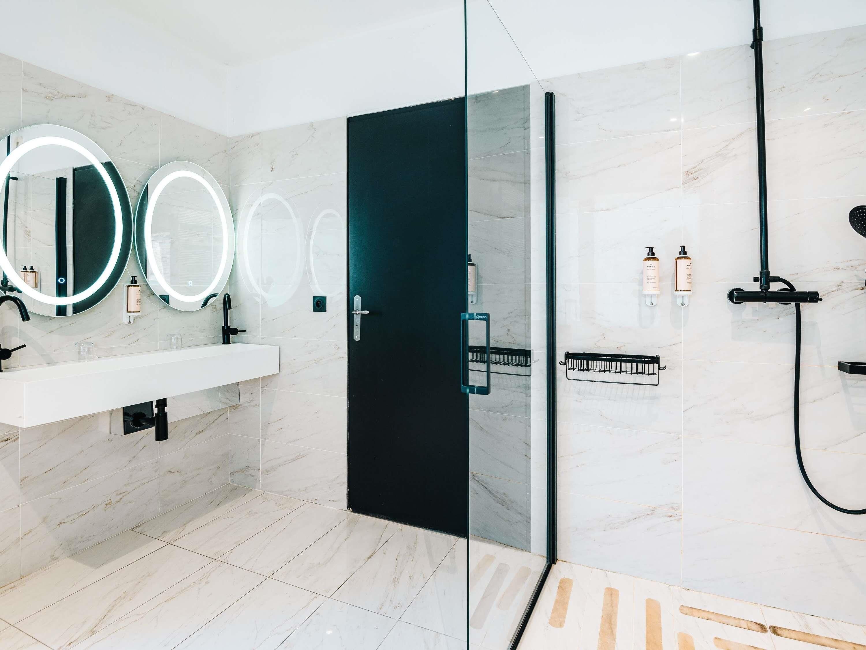 A modern bathroom with a glass shower enclosure, a black door, and three illuminated mirrors.