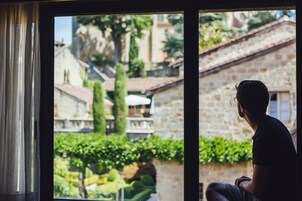 Una persona sentada junto a una ventana con vista a un patio con edificios de piedra y vegetación.