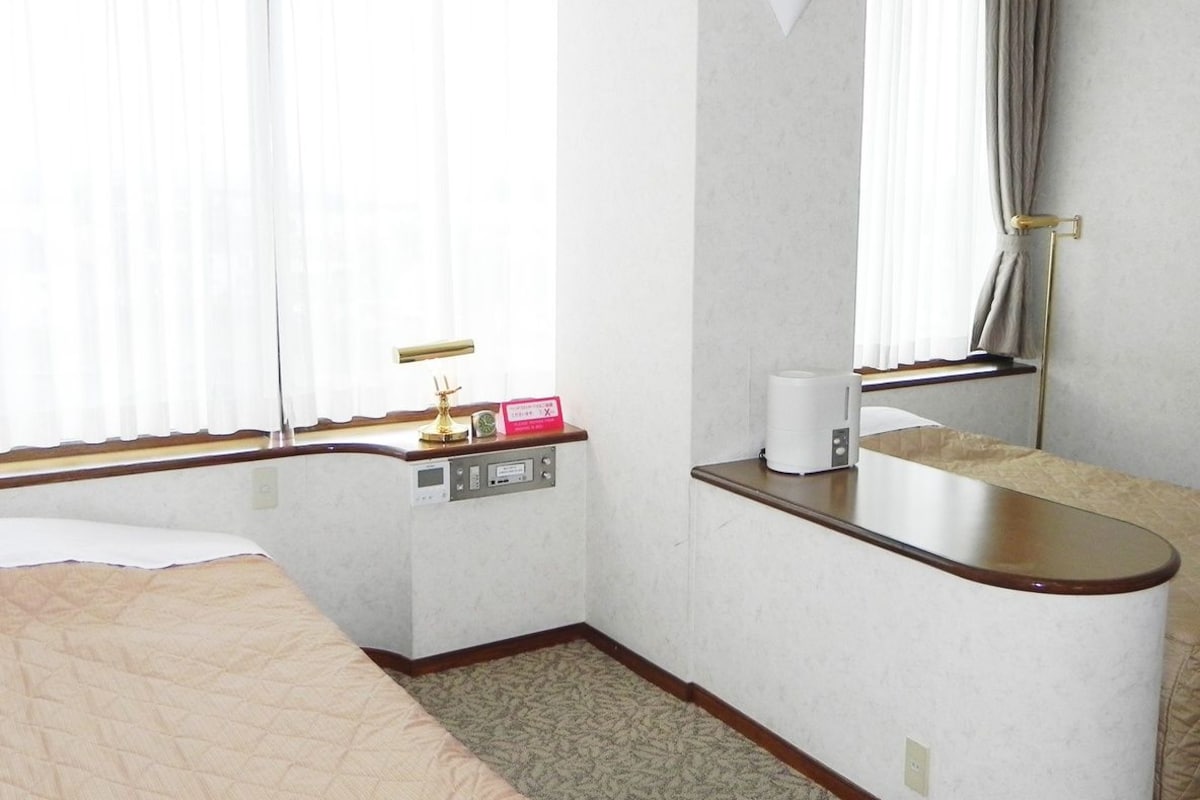 A hotel room with a bed, a desk with a chair, a mirror, and a window with curtains.