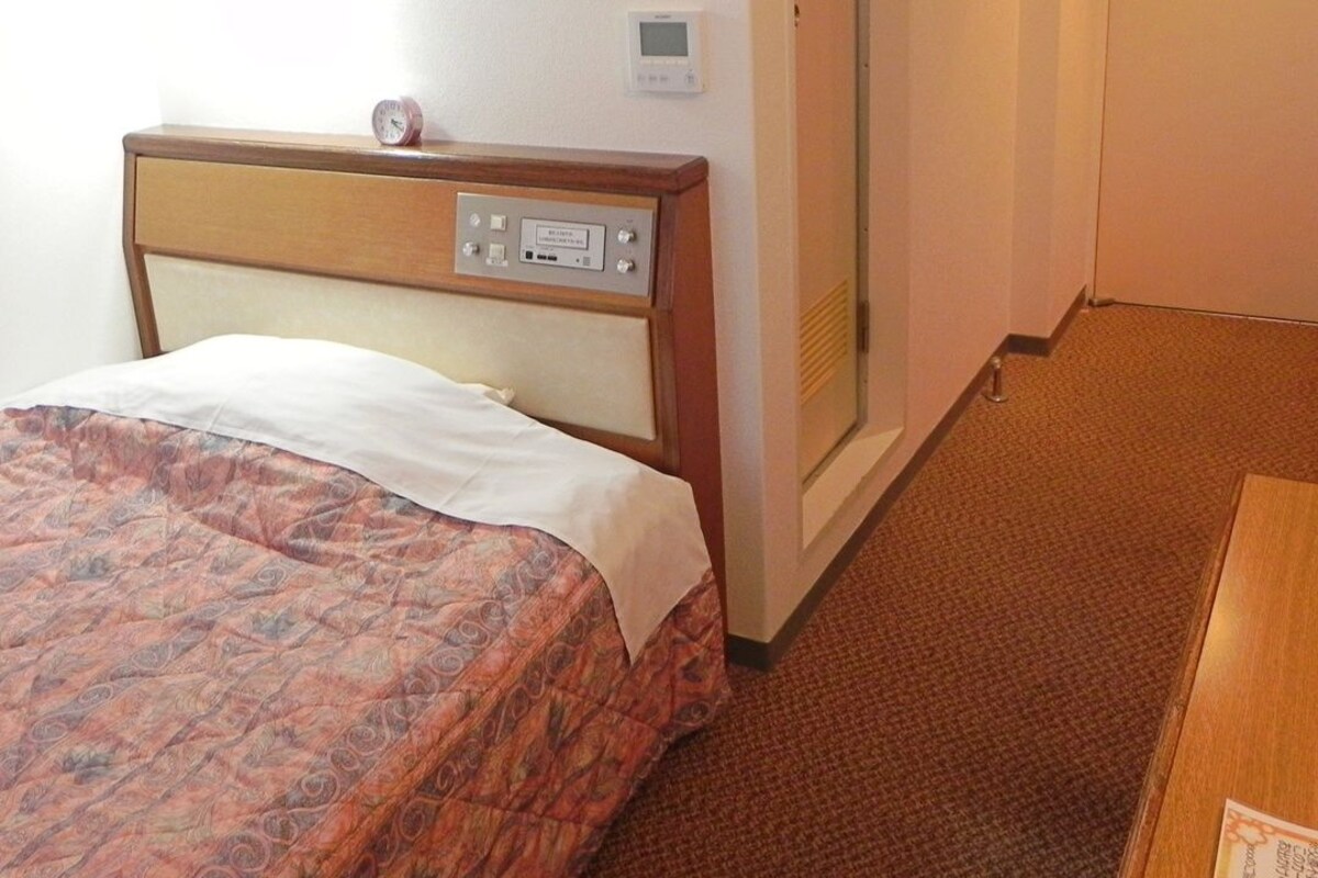 A hotel room with a bed, a bedside table, a clock, and a thermostat on the wall.