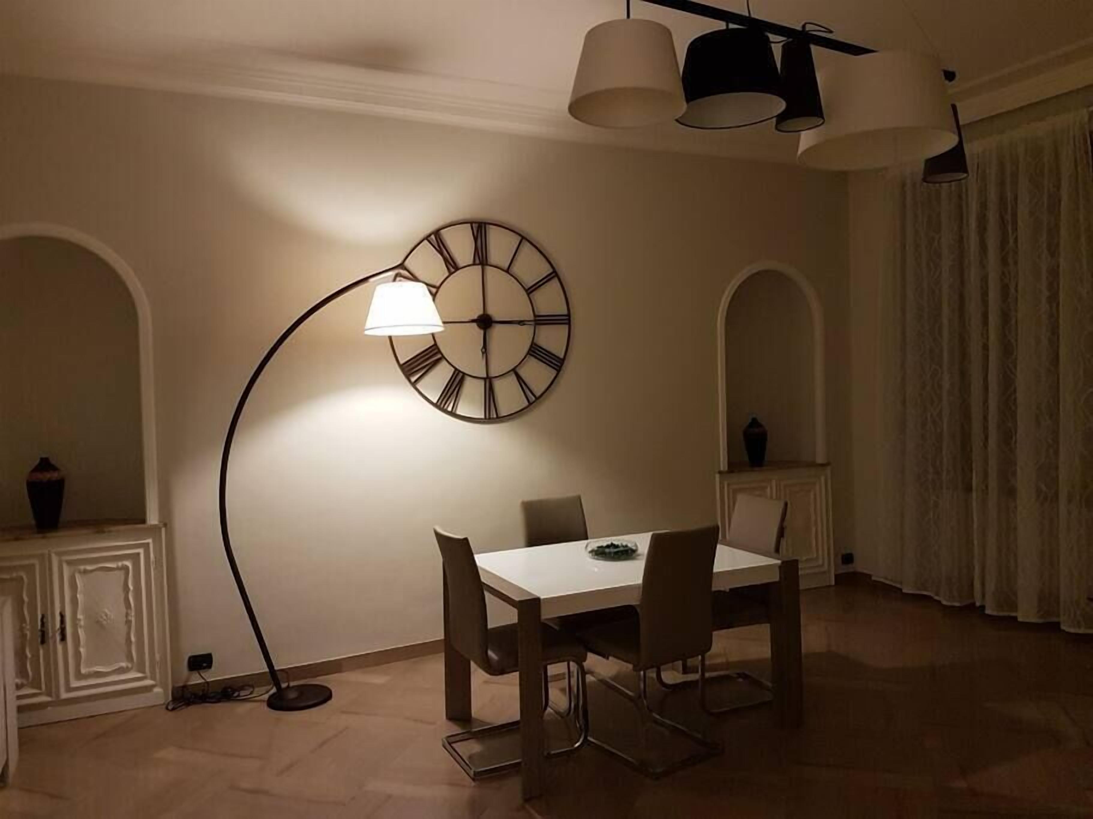 A dining area with a white table, four chairs, a wall-mounted clock, and a floor lamp.