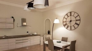 A modern kitchen with a dining area, featuring a large wall clock, pendant lights, and a minimalist design.