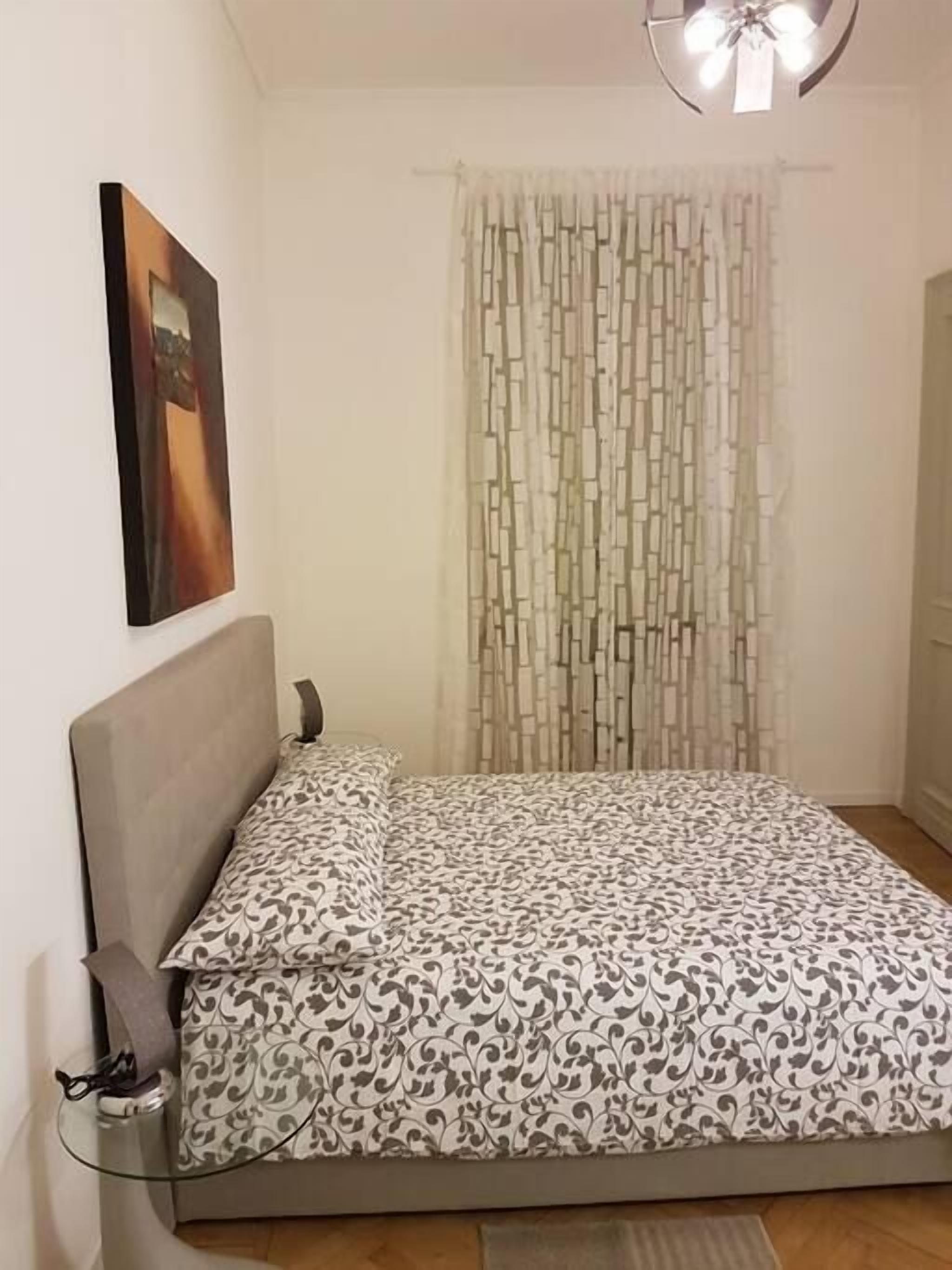 Une chambre à coucher avec un lit, une table de chevet avec une lampe, un tableau accroché au mur et un rideau orné d’un motif géométrique.