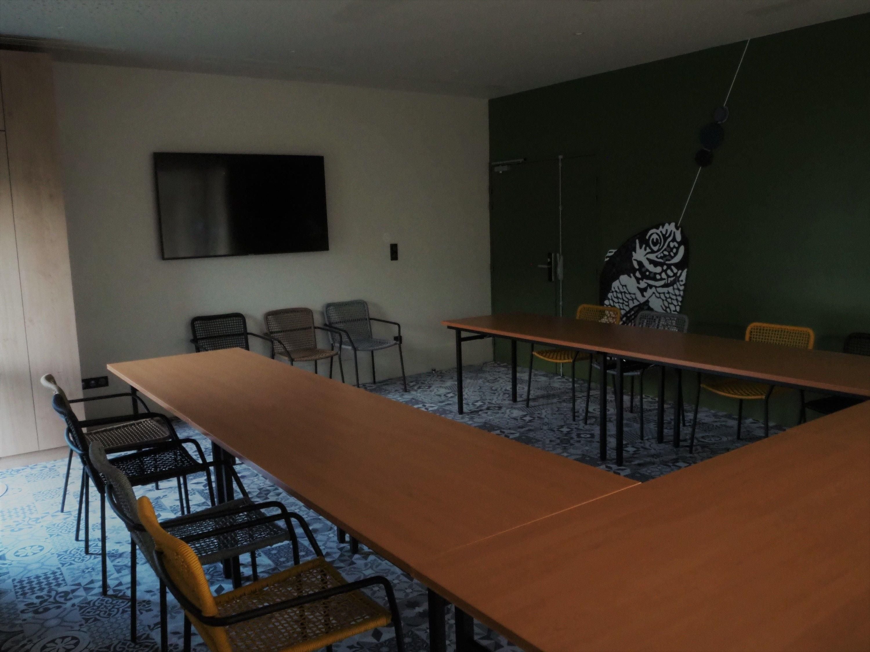 A conference room with a long table, several chairs, a flat-screen TV on the wall, and a patterned carpet.