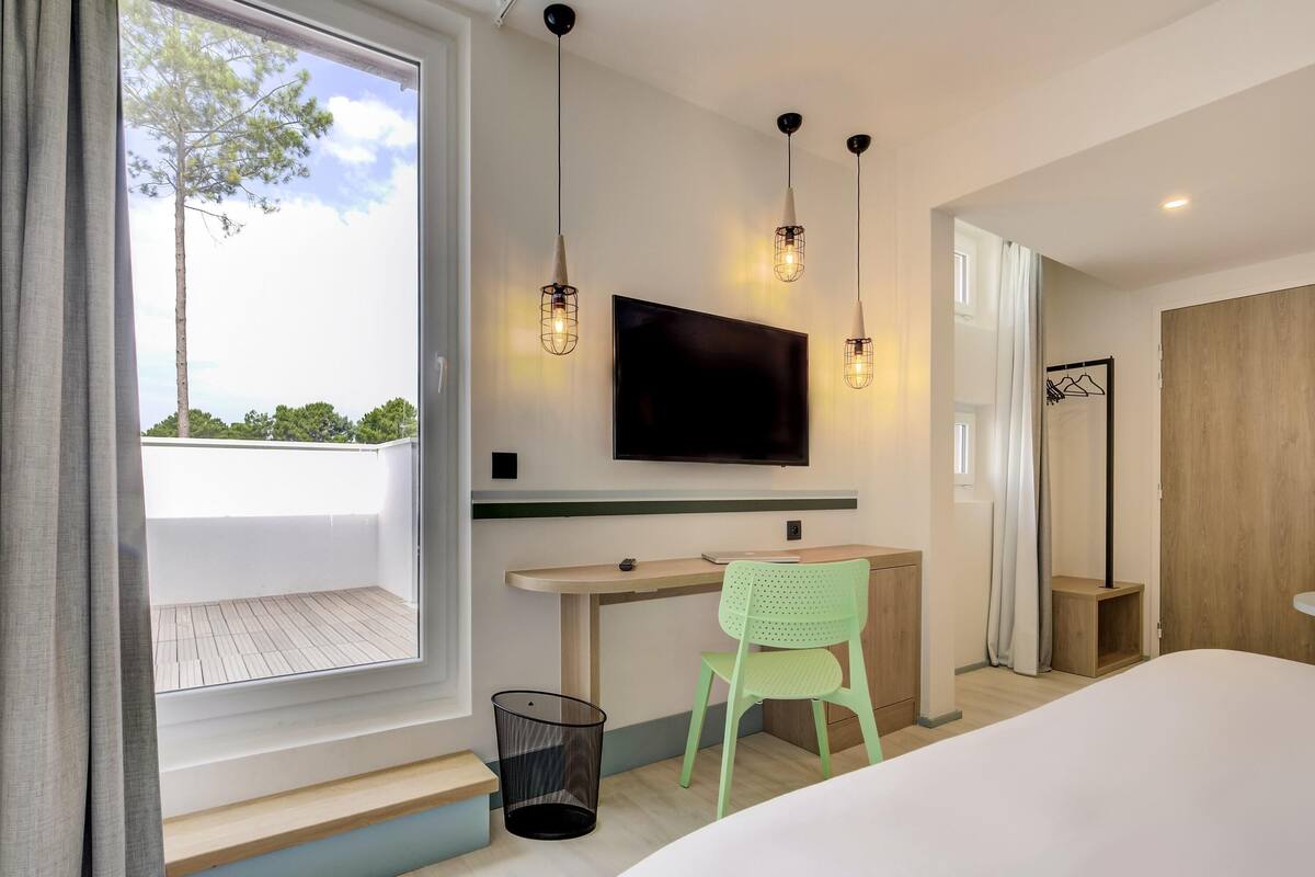 Une chambre d’hôtel moderne dotée d’un grand lit, d’un bureau avec une chaise, d’un téléviseur fixé au mur et d’une fenêtre donnant sur la verdure.
