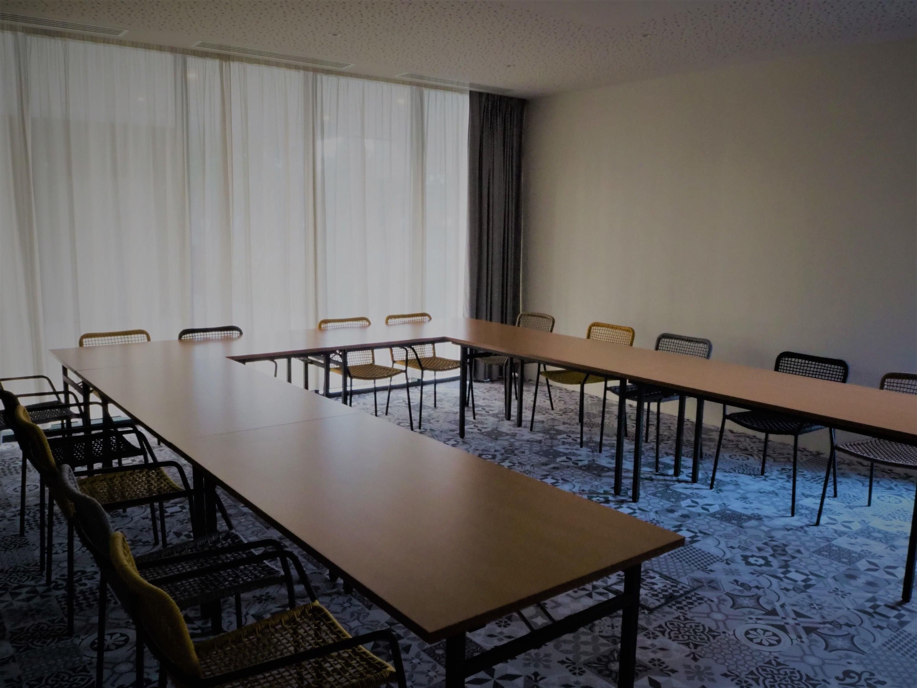 Une salle de conférence avec une longue table et des chaises, de grandes fenêtres avec des rideaux et un tapis à motifs.
