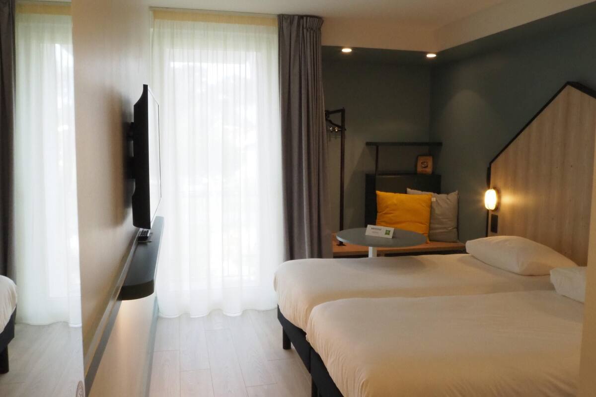 Une chambre d’hôtel avec deux lits, un bureau, une chaise, une télévision et une fenêtre avec des rideaux.