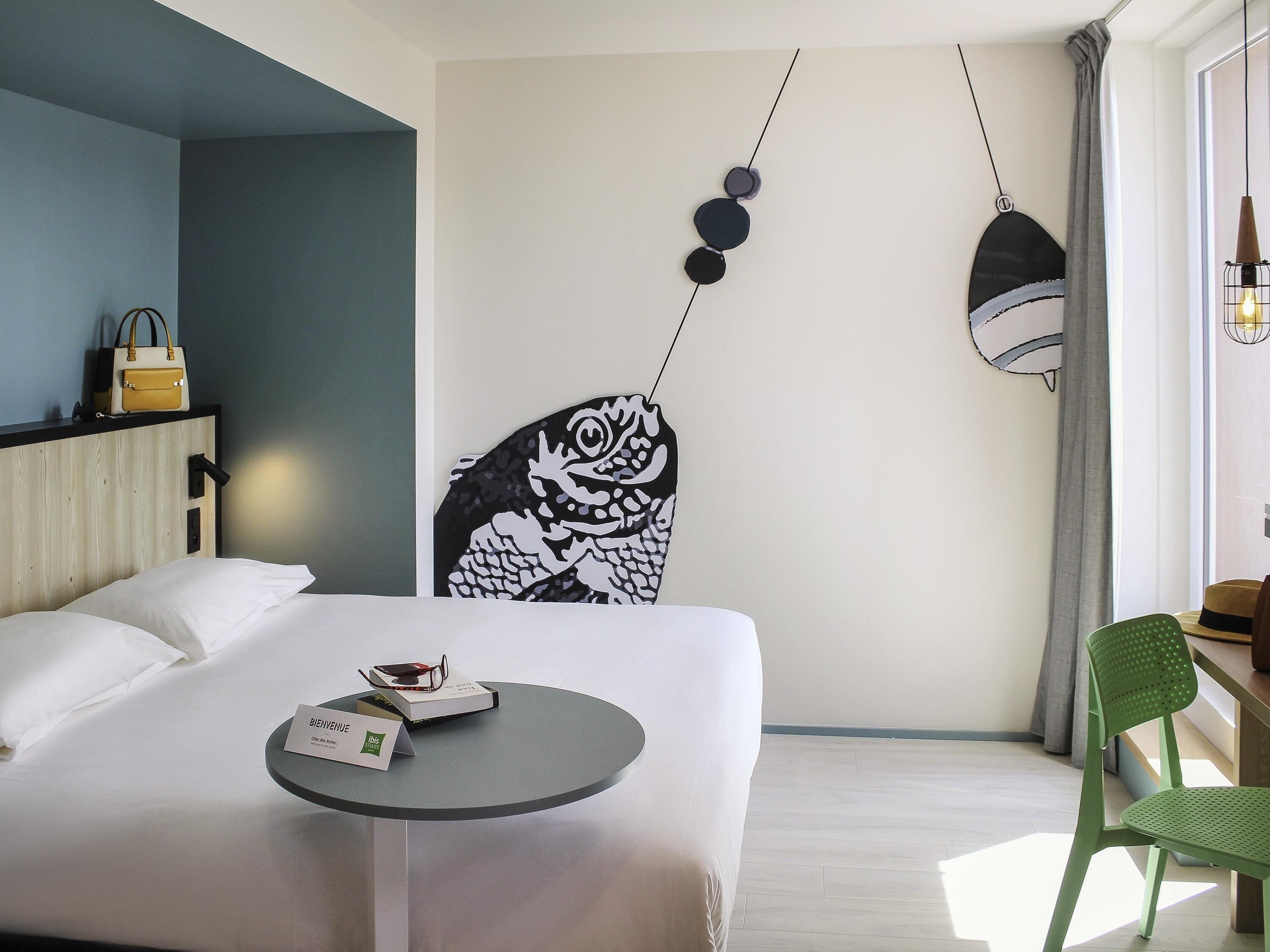 A hotel room with a bed, a small round table, a green chair, a wall-mounted artwork, and a hanging light fixture.