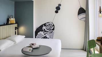 A hotel room with a bed, a small round table, a green chair, a wall-mounted artwork, and a hanging light fixture.