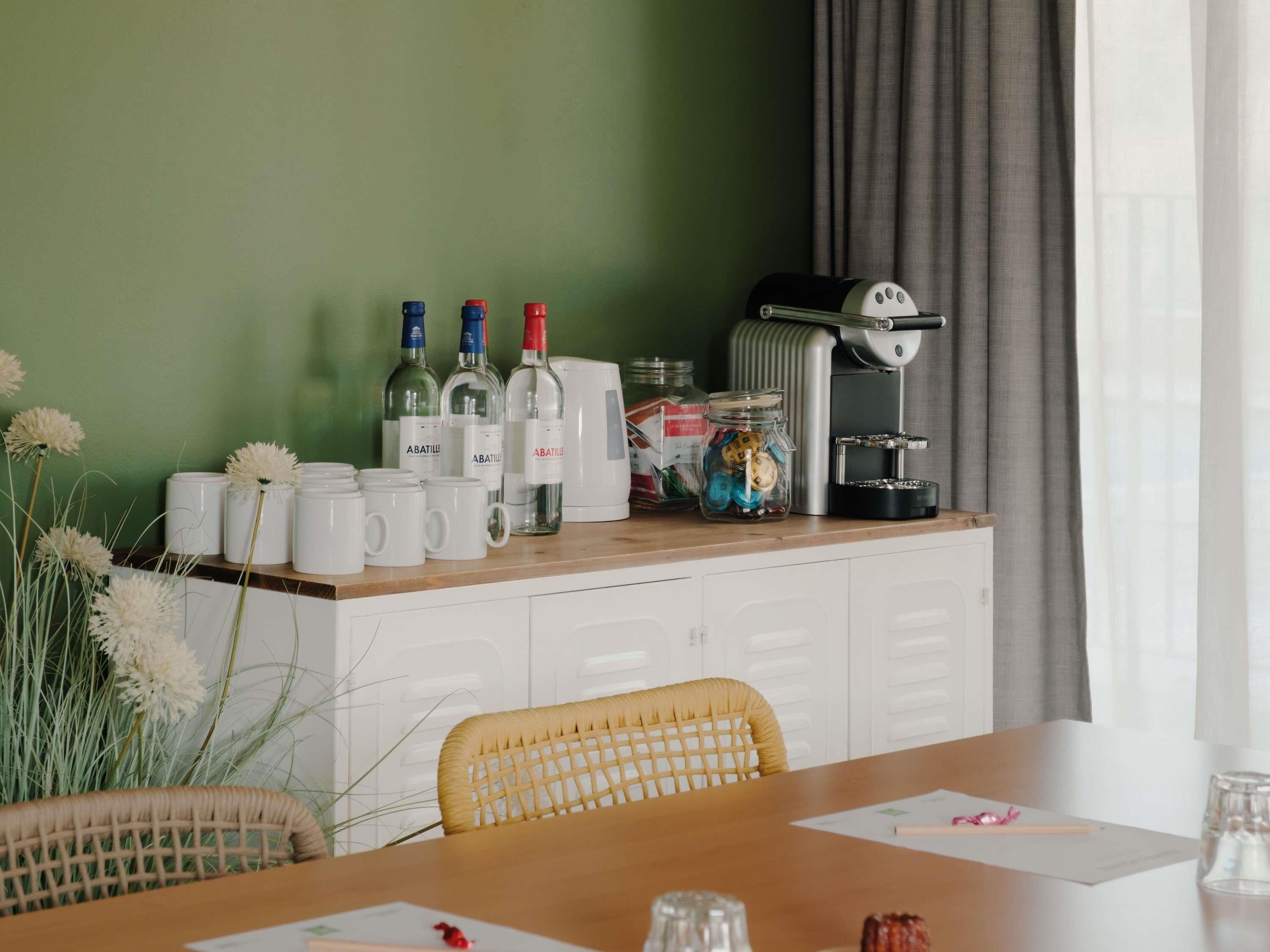 Quarto de hotel com mesa de jantar, armários brancos, máquina de café e objetos decorativos.