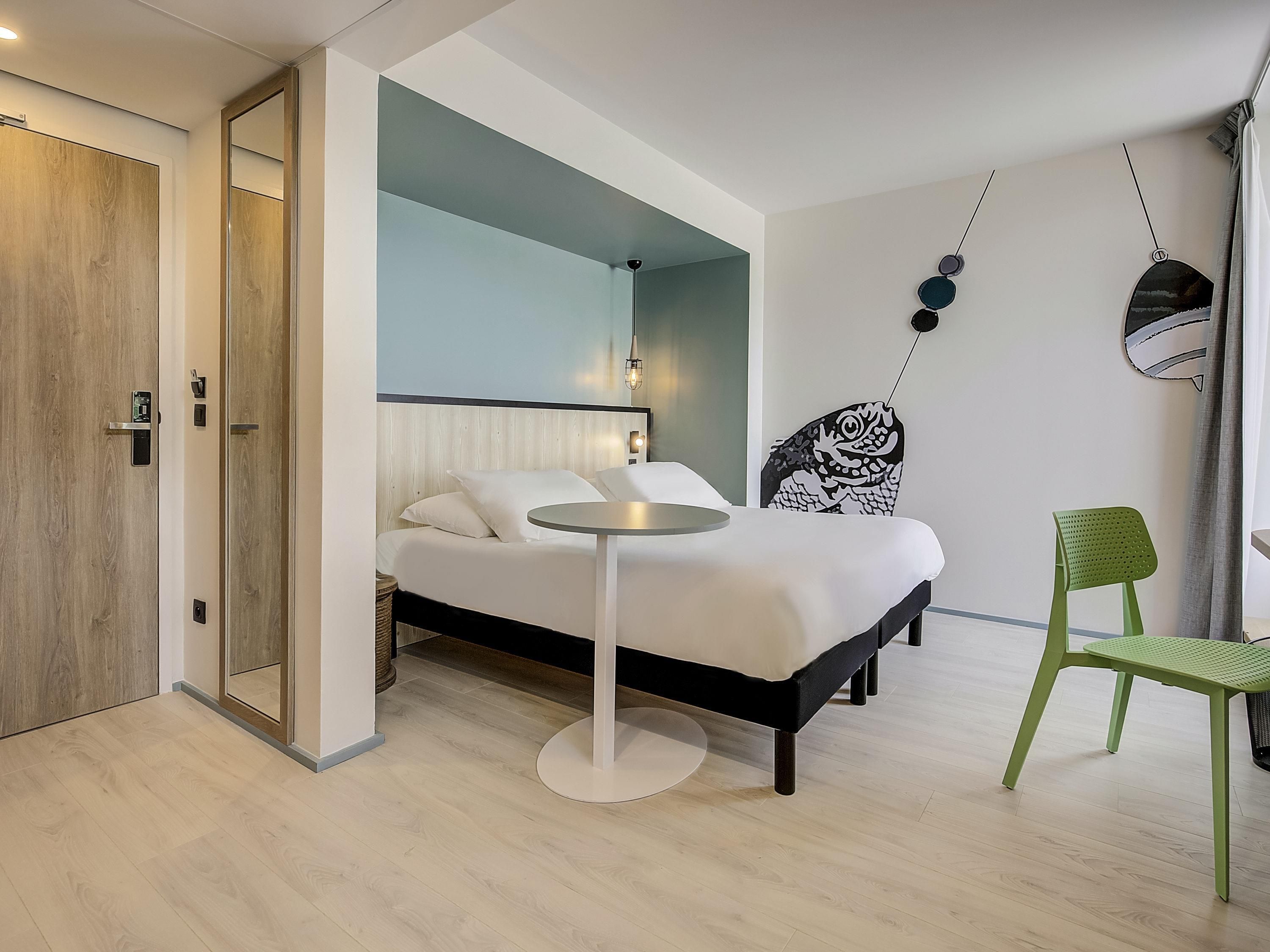 A modern hotel room with a bed, a round table, a green chair, and a mirror on the wall.
