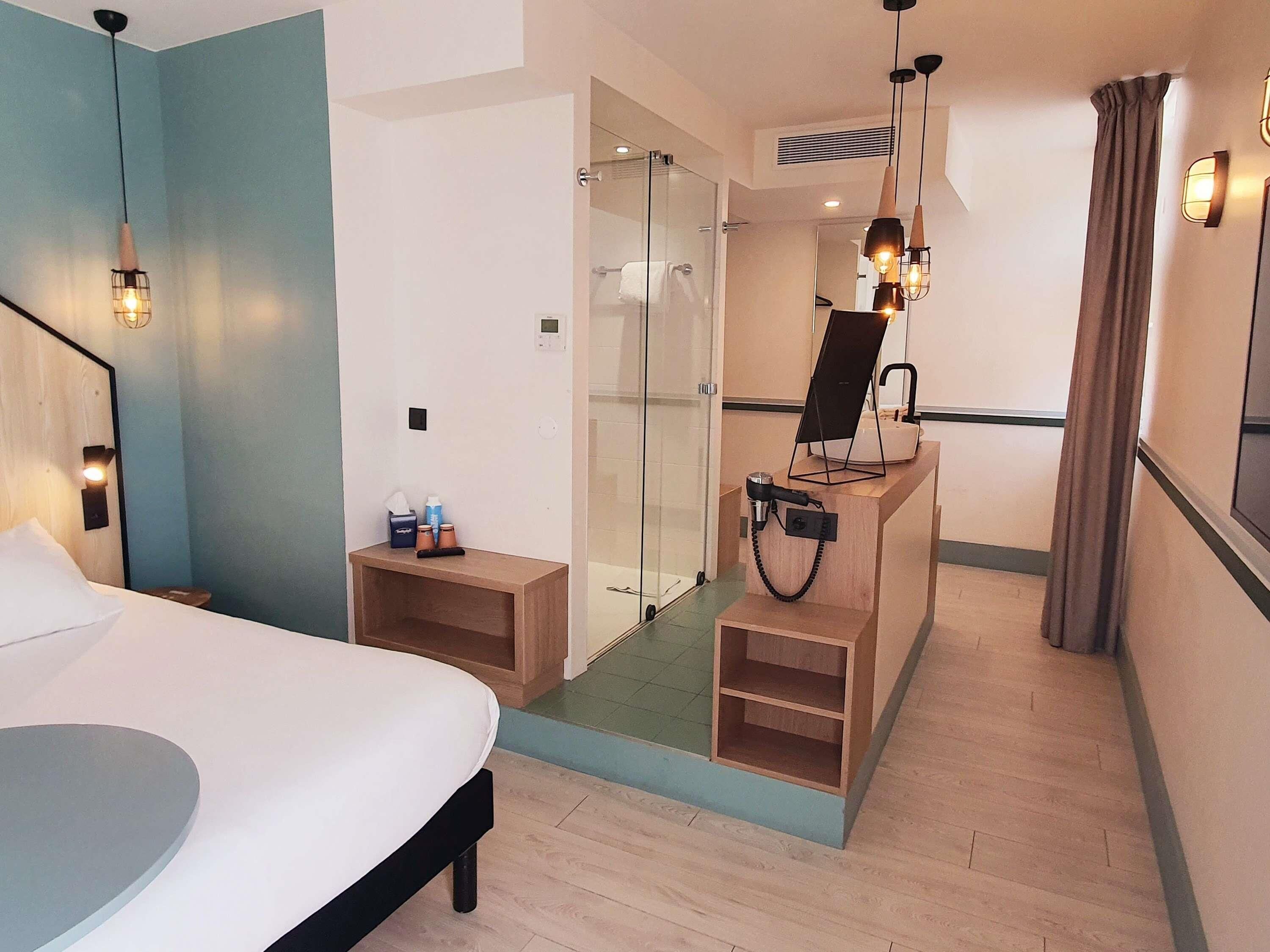 Habitación de hotel con cama, mesita de noche, espejo, ducha y un pequeño armario de madera.