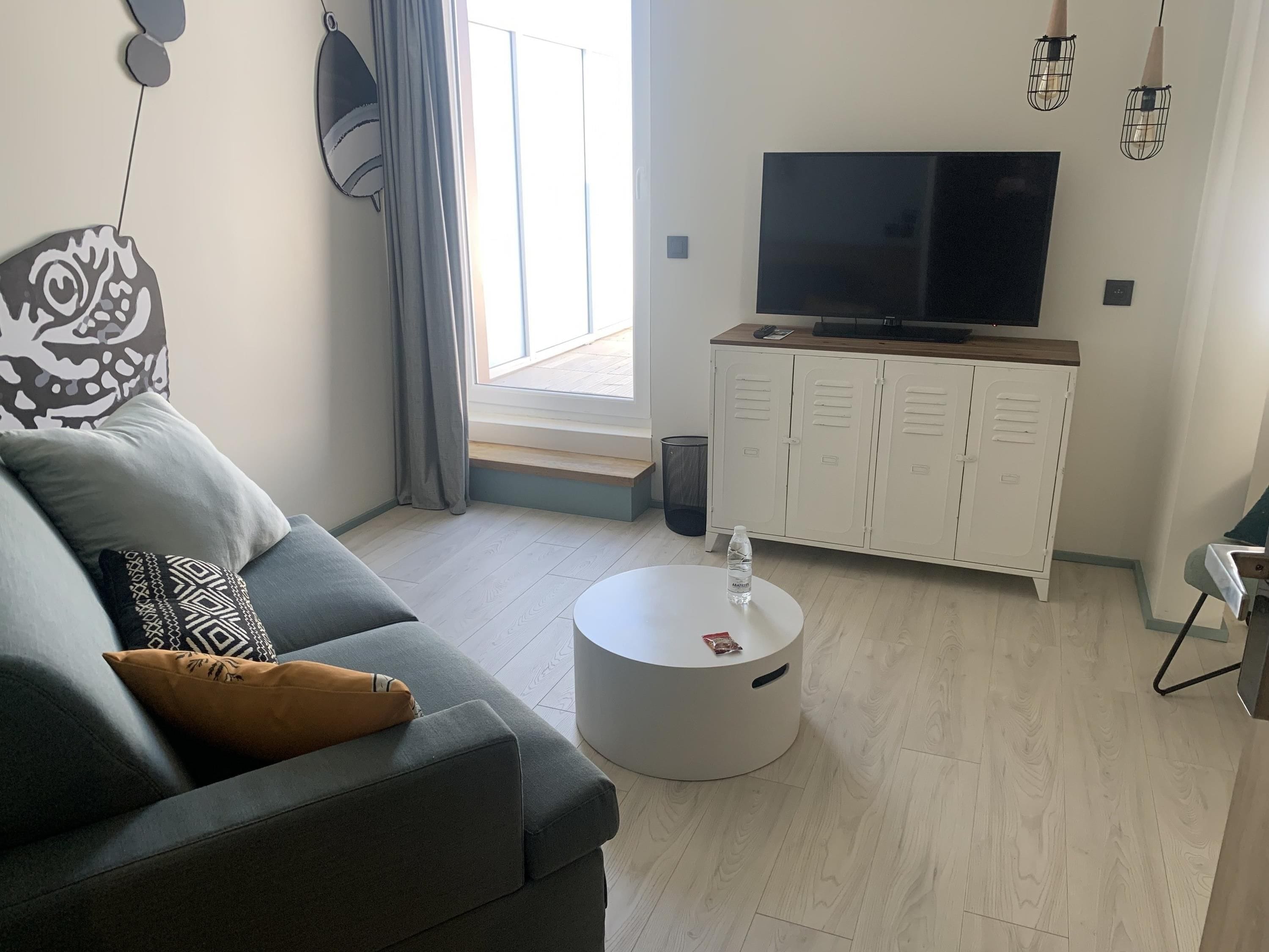 Un salon moderne avec un canapé gris, une table basse ronde, un téléviseur posé sur un meuble en bois et une fenêtre avec des rideaux.
