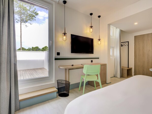 A modern hotel room with a large bed, a desk with a chair, a TV mounted on the wall, and a window with a view of trees.