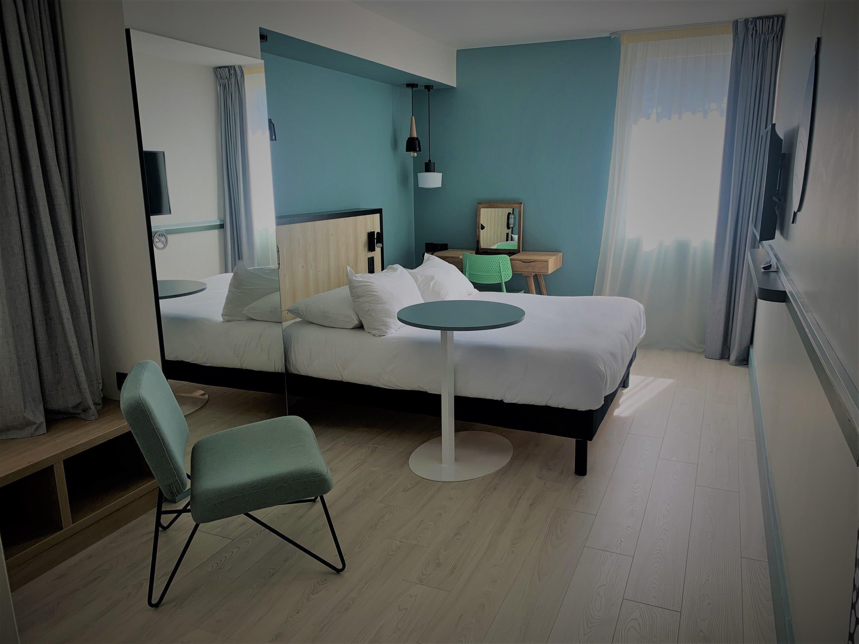 Ein Hotelzimmer mit einem Bett, einem runden Tisch, einem Stuhl, einem Schreibtisch, einem Spiegel und einem Fenster mit Vorhängen.