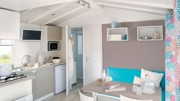A compact kitchen with a microwave, stove, and sink. There is a dining area with a table and chairs, and a blue sofa. Shelves with decorative items are on the wall. - Camping Les Tilleuls - Mobilhome (Rocamadour)