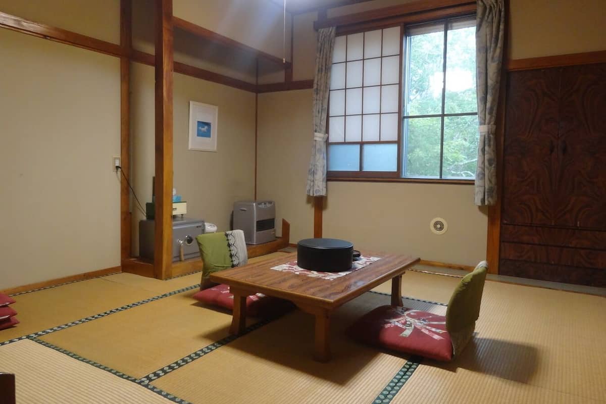 Um quarto japonês tradicional com tatames, uma mesa de madeira, uma cadeira verde, uma janela com vista e uma porta deslizante.
