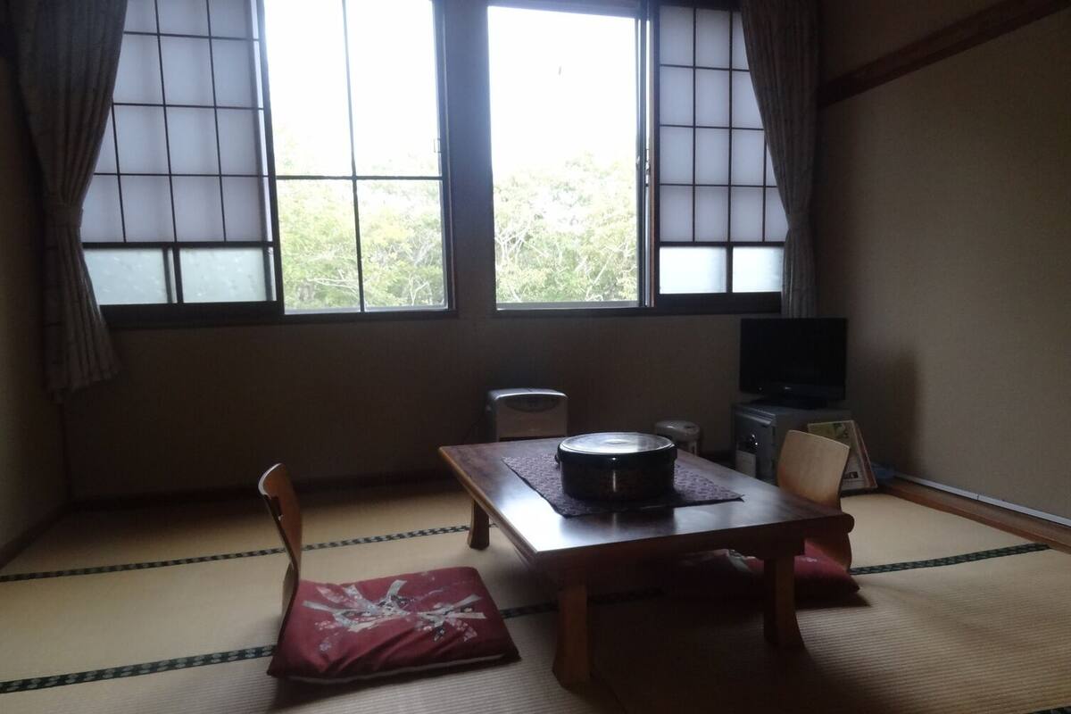 Um quarto japonês tradicional com tatames, uma mesa baixa com almofada, uma televisão e uma janela com cortinas.