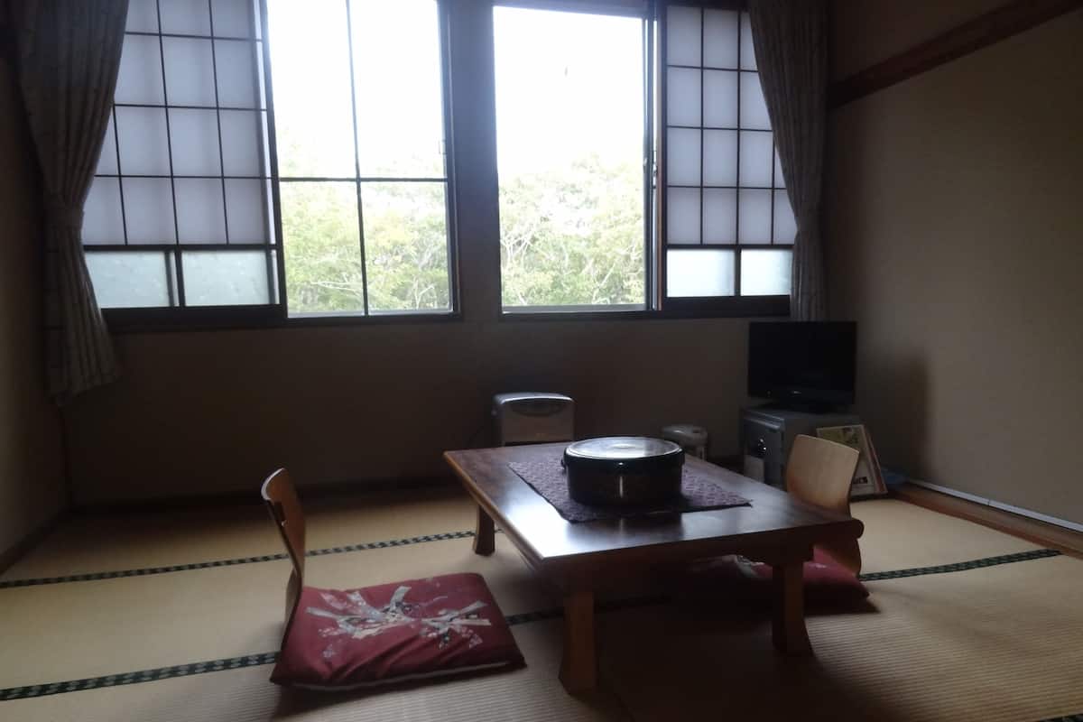 Um quarto japonês tradicional com tatames, uma mesa baixa, um assento de almofada, uma televisão e uma janela com vista para árvores.