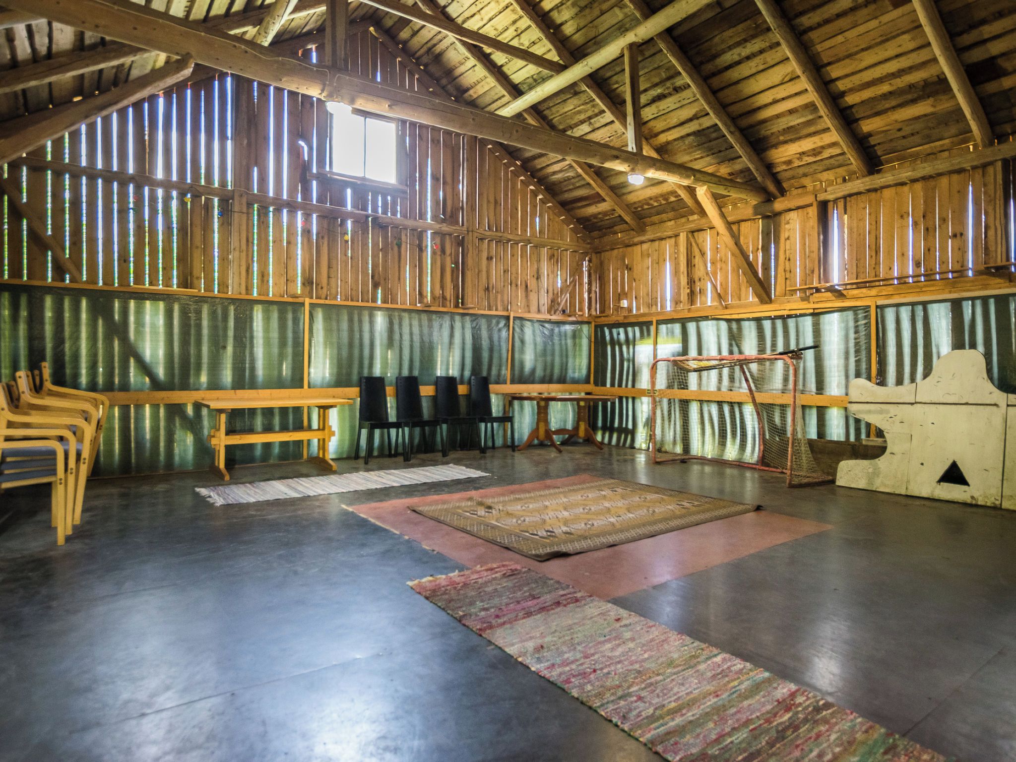 Interior rústico de un granero con vigas de madera, ventanas con marco de metal y un amplio espacio abierto con un banco, sillas y una alfombra estampada.