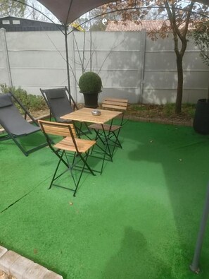 Een terras buiten met kunstgras, een houten tafel met stoelen en een plant in pot.