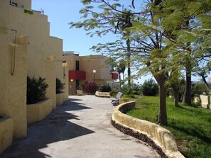 A pathway leading to a building with a red balcony, surrounded by greenery and a tree.