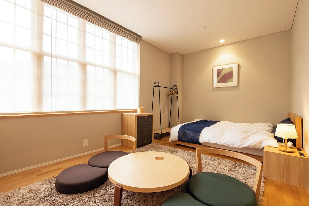 A bedroom with a bed, bedside table, lamp, chair, round table, and a window with blinds.