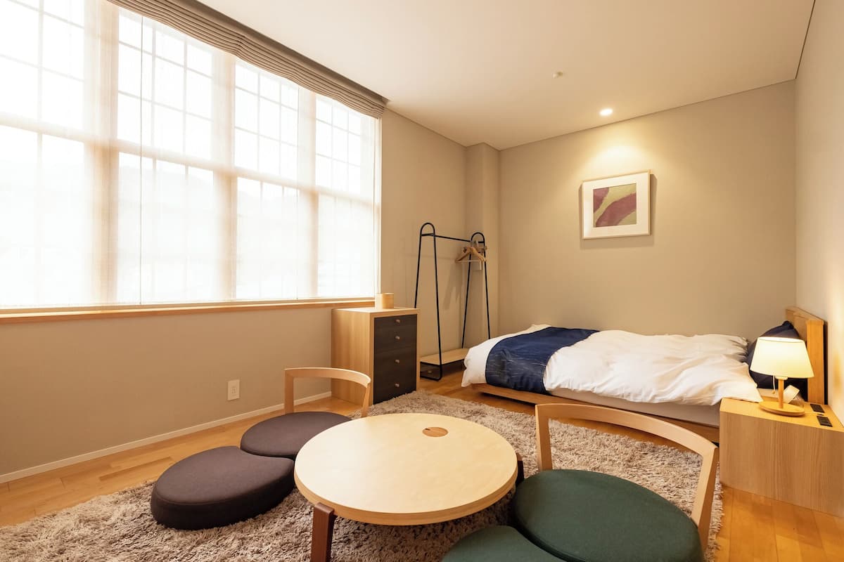 Ein Schlafzimmer mit Bett, Nachttisch, Lampe, Stuhl, rundem Tisch und einem Fenster mit Jalousien.