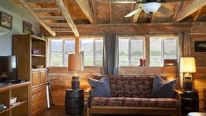 Et hyggeligt sommerhusindretning med træloft, sofa, fjernsyn og et stort vindue med udsigt til naturen.