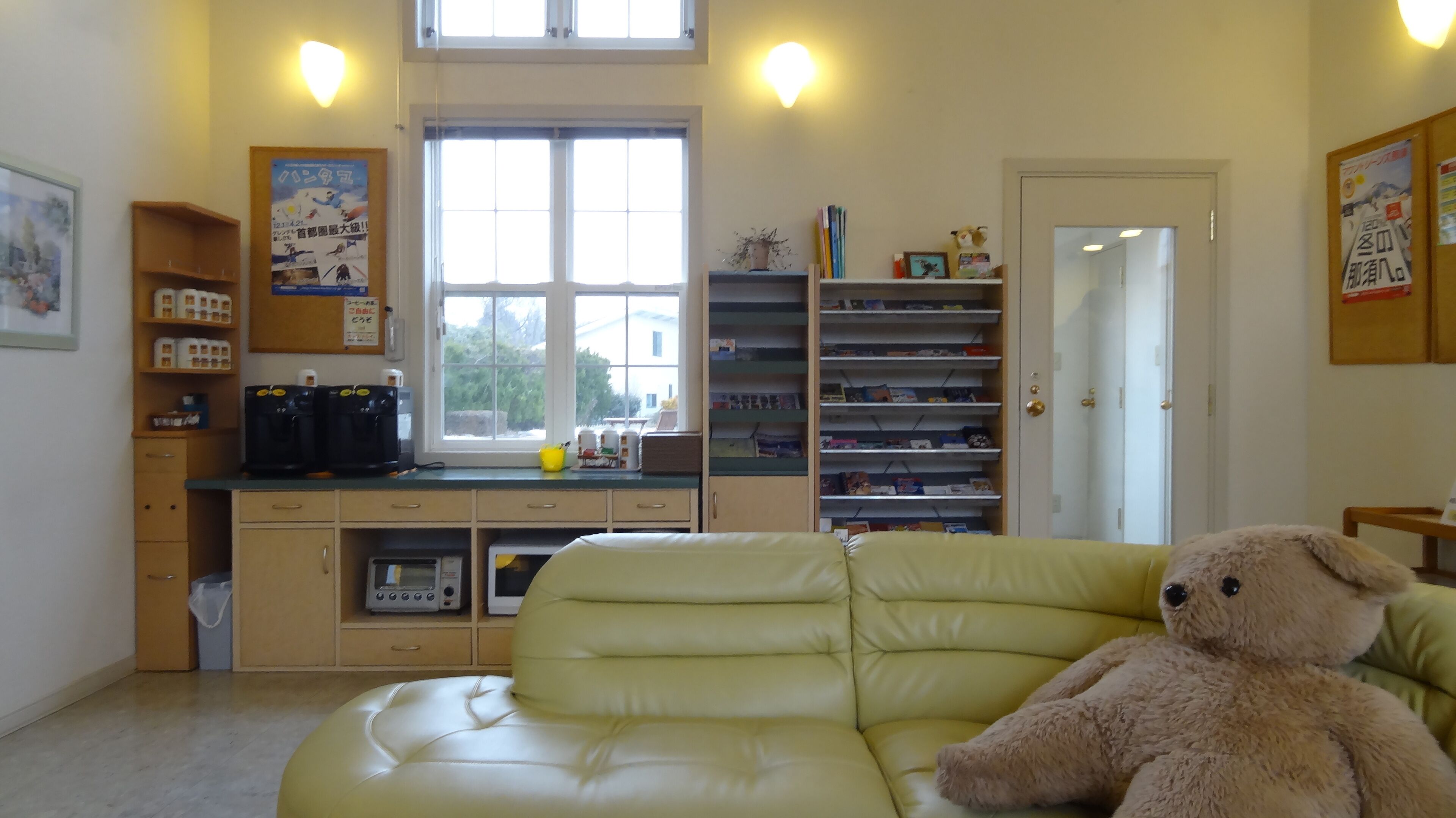 A room with a green leather sofa, a teddy bear, a coffee machine, and a window with a view of greenery.