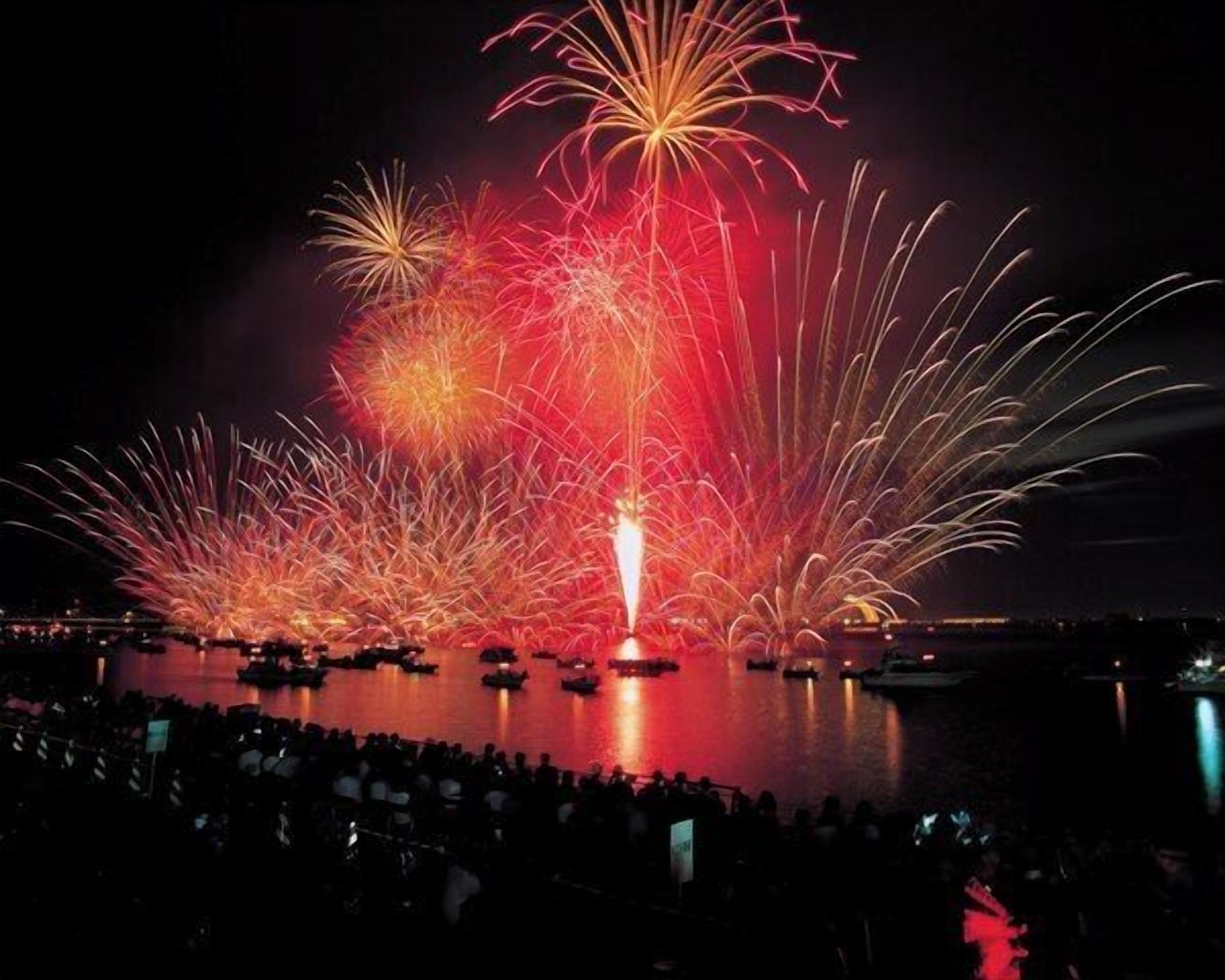 A nighttime fireworks display over a body of water with boats and a crowd of spectators.