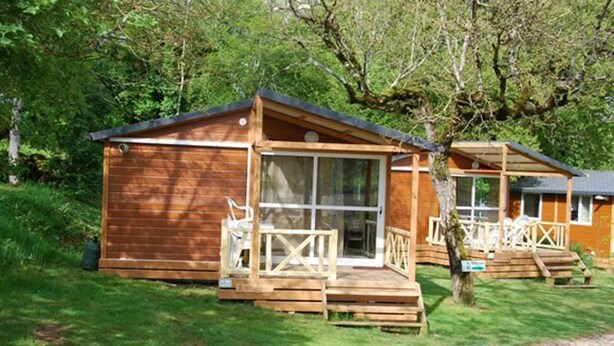 A wooden cabin with a porch and a glass door, surrounded by trees. - Camping Les Tilleuls - Chalets (Rocamadour)