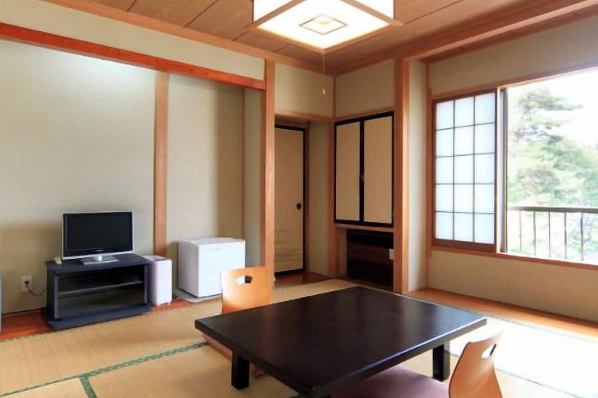 Una habitación japonesa tradicional con suelo de tatami, una mesa baja, un televisor sobre un soporte y puertas correderas.