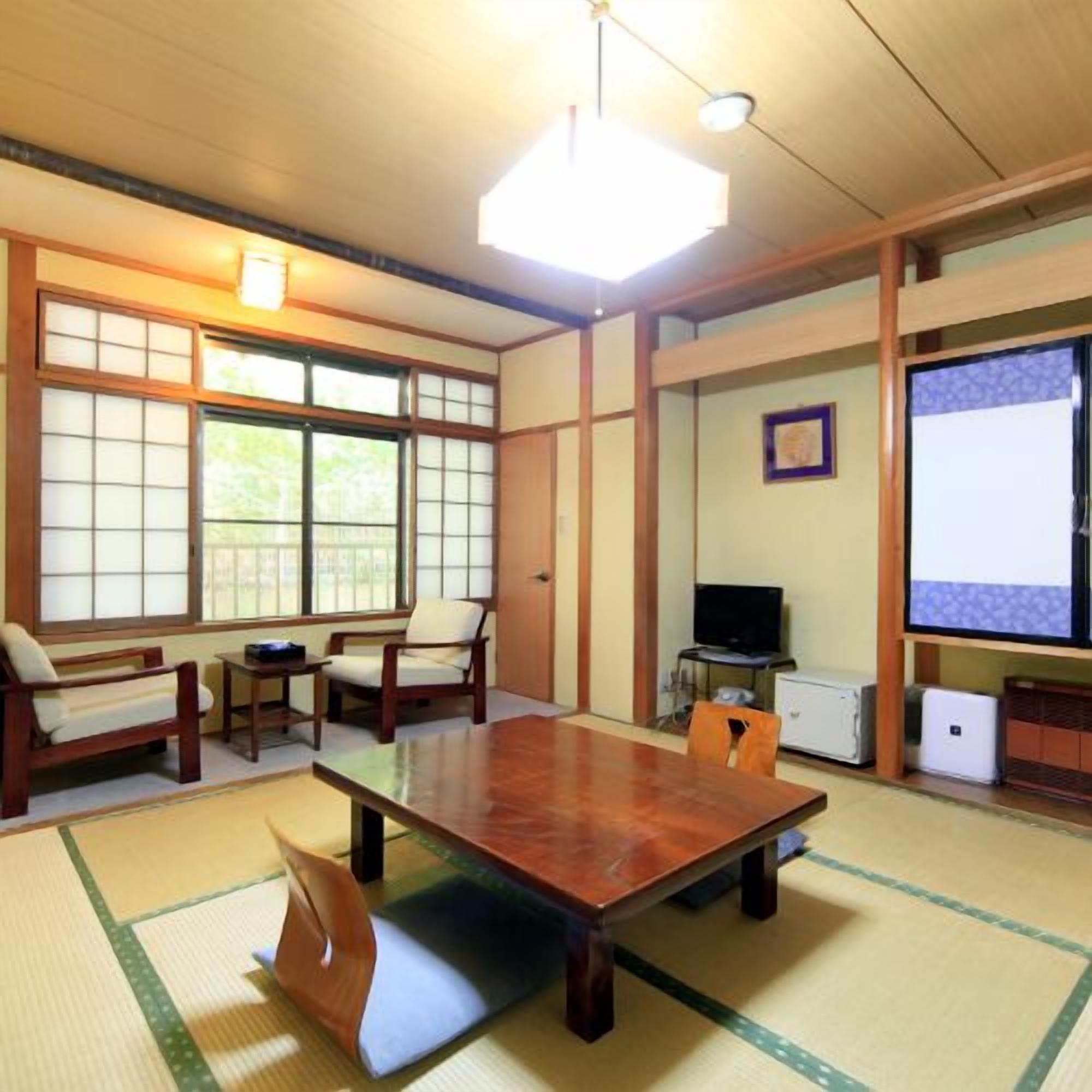 A traditional Japanese room with tatami flooring, a low table, wooden chairs, a television, and a projector screen.