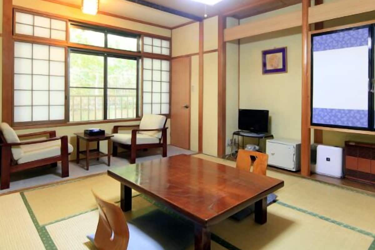 Una habitación japonesa tradicional con suelo de tatami, una mesa baja, sillas de madera, un televisor y una pantalla de proyector.