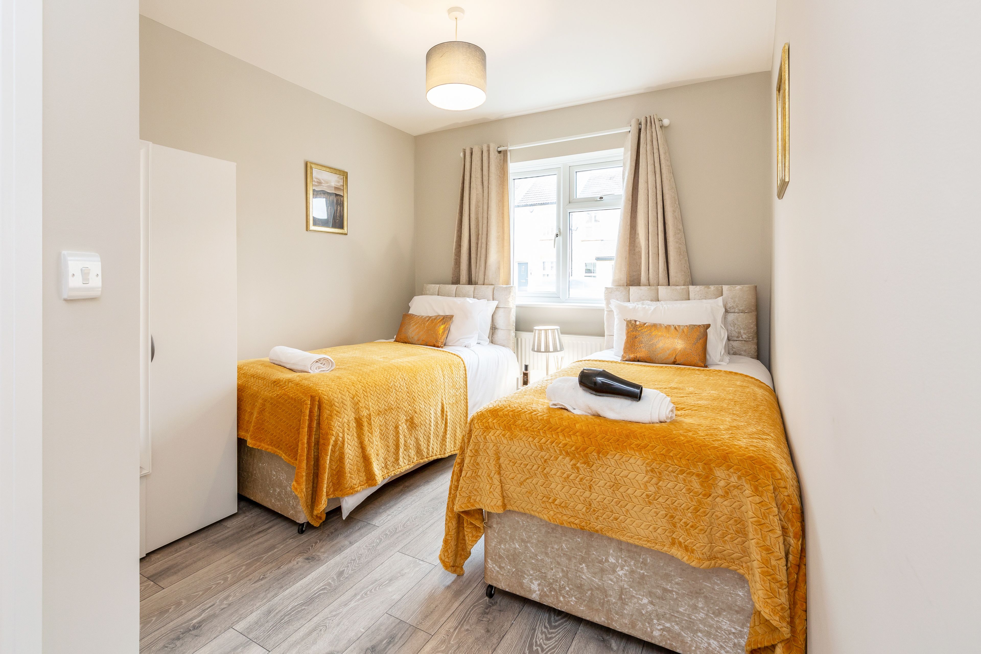 A room with two beds, each with a yellow blanket, a white door, a window with curtains, and a ceiling light.