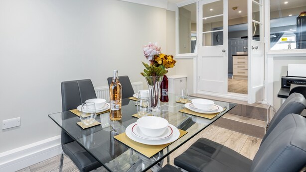A modern dining area with a glass table, black chairs, and a vase of flowers. - HiikotsHomes - Dartford (Dartford)
