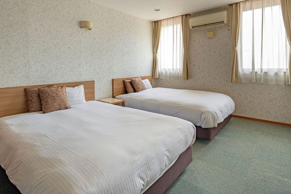 A hotel room with two beds, a small bedside table, a wall-mounted air conditioner, and a patterned wallpaper.
