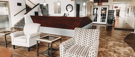 Un área de recepción con un mostrador de madera, un sillón blanco, un sillón estampado y una mesa con cubierta de vidrio.