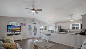 A spacious living room with a television, a coffee table, and a sofa. There is a kitchen area with white cabinets and a dining area with white chairs.