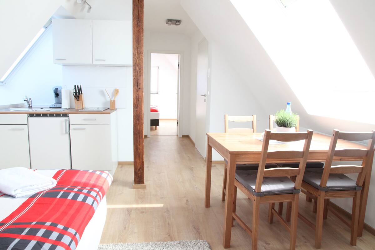 Quarto moderno e bem iluminado com uma mesa de jantar de madeira, quatro cadeiras e uma cama com colcha xadrez vermelha e preta.