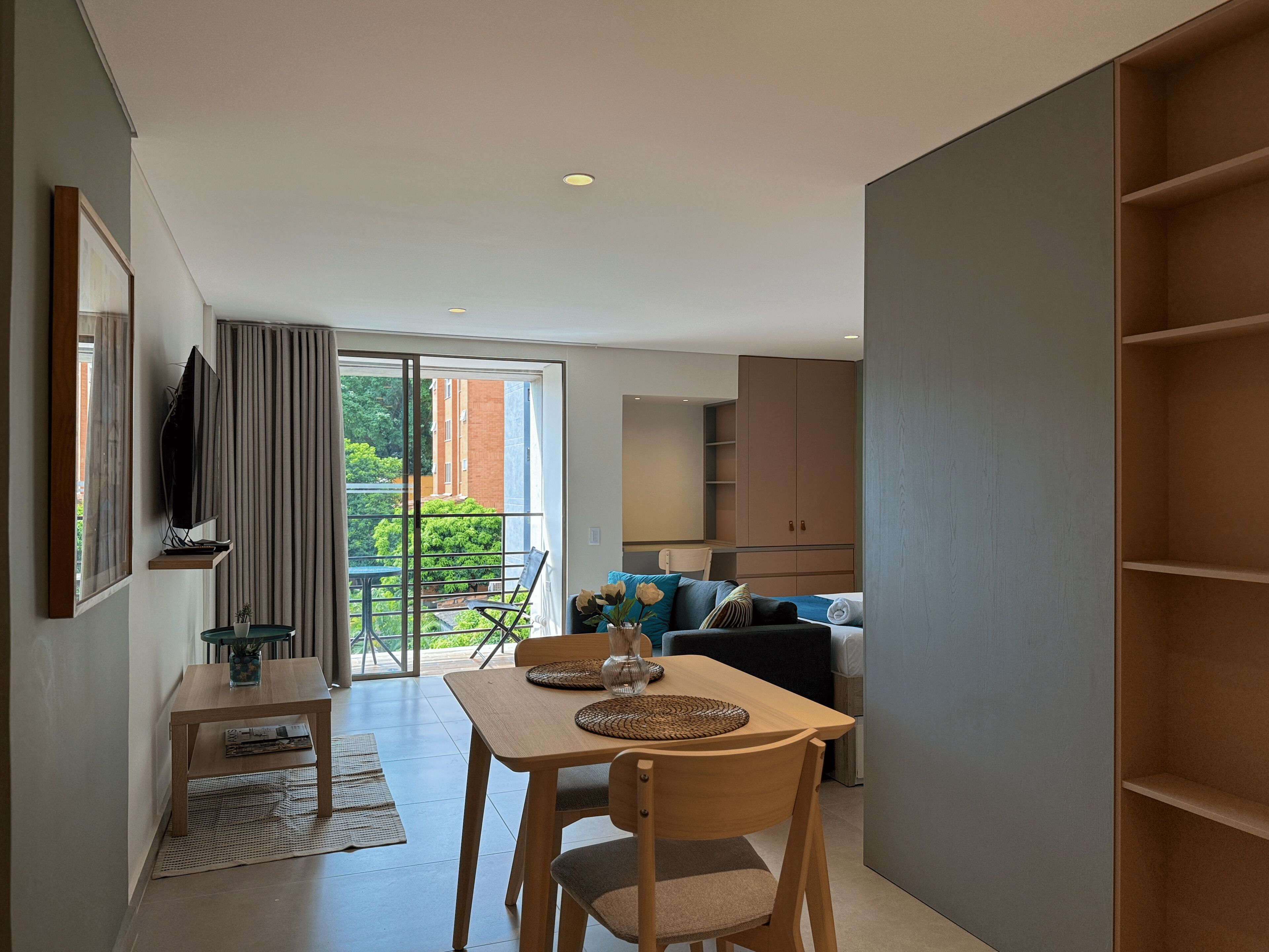 A modern living space with a dining area, a sofa, a TV, and a balcony with a view of greenery.
