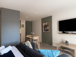 A modern hotel room with a sofa, a small dining table with chairs, a flat-screen TV mounted on the wall, and a framed artwork above the TV.