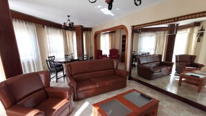 Un salón amplio con un sofá de cuero marrón, un sillón a juego y una mesa de centro de madera. Se ve un rincón de comedor con una mesa y sillas al fondo.