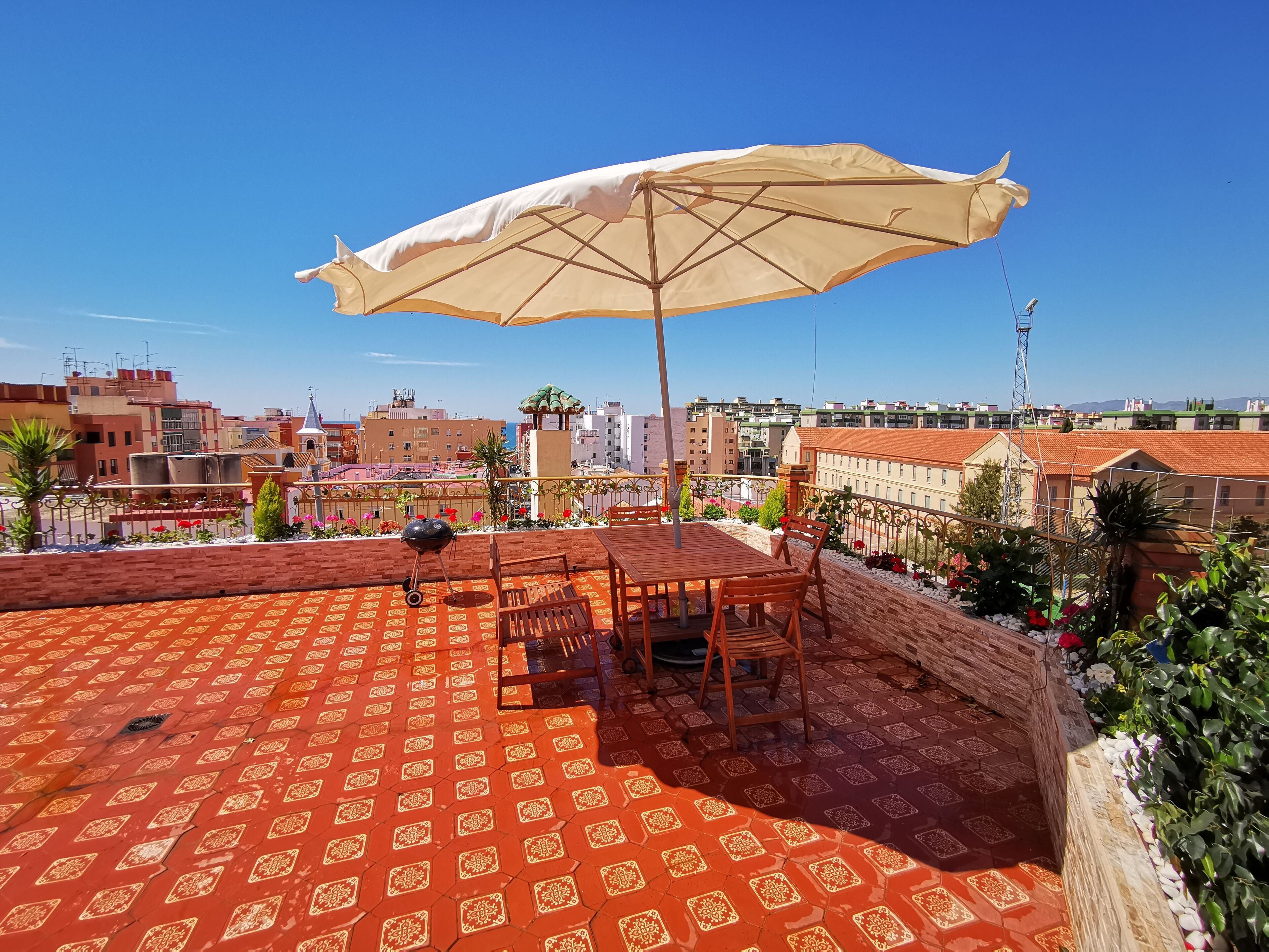 A rooftop terrace with a patterned red floor, a wooden table and chairs, a barbecue grill, and a large beige umbrella.